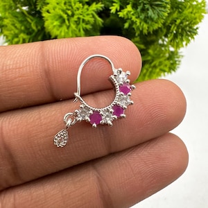 Puede incluir: Un aro de nariz plateado con una disposición semicircular de piedras redondas alternas en rosa y transparente. La pieza de joyería incluye un pequeño dije colgante en forma de lágrima incrustado con piedras transparentes en el lateral.