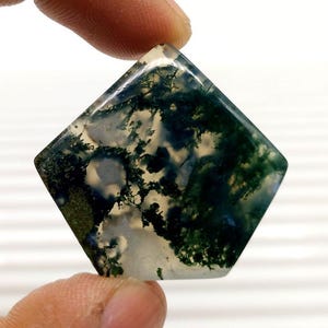 Pode incluir: Uma pedra preciosa de corte quadrado com um padrão de ágata musgo. A pedra apresenta uma base translúcida com inclusões verdes, brancas e pretas, criando um design natural. A gema é segurada entre dois dedos.