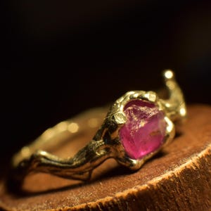 May include: A gold-colored ring featuring a raw, pink gemstone. The ring has a textured, organic design, with the gemstone held in a claw-like setting. The ring is displayed on a wooden surface against a dark background.