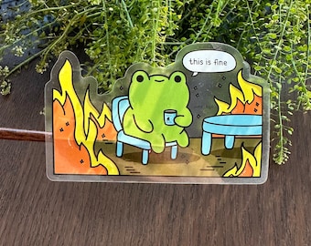 Op de afbeelding: Een doorzichtige sticker met een cartoonkikker die in een stoel zit, een mok vasthoudt, omringd door vlammen. De tekstballon zegt "this is fine". De sticker heeft een humoristisch en herkenbaar thema.