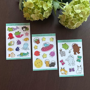 Puede incluir: Tres hojas de pegatinas con ilustraciones coloridas de críptidos y elementos espaciales. Las pegatinas presentan varias criaturas, un OVNI, planetas, estrellas y plantas. El texto "cryptids with plants and friends" está impreso en cada hoja.