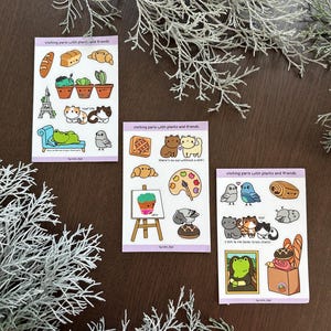 May include: Three sticker sheets with illustrations of cats, birds, plants, and pastries. The stickers are on a light purple background with the text "visiting paris with plants and friends".