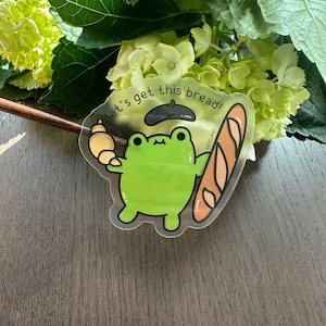 Puede incluir: Una rana verde con un gorro negro sostiene una baguette y un croissant. La rana está en una pegatina transparente con el texto "let's get this bread!" y "cute_togo".