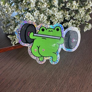 Può includere: Un adesivo olografico con un simpatico ranocchio verde che solleva un bilanciere. Il ranocchio sorride e solleva pesi. L'adesivo ha un bordo glitterato e il testo "cute_kojo" è visibile.