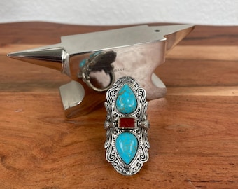 May include: An ornate silver ring with two turquoise teardrop-shaped stones and a rectangular red stone in the center. The ring has intricate filigree detailing and sits on a wooden surface.