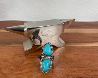 May include: A silver-toned ring featuring two turquoise-colored stones. The ring has an ornate design with a detailed setting. The ring is displayed on a wooden surface with a silver anvil-shaped object in the background.