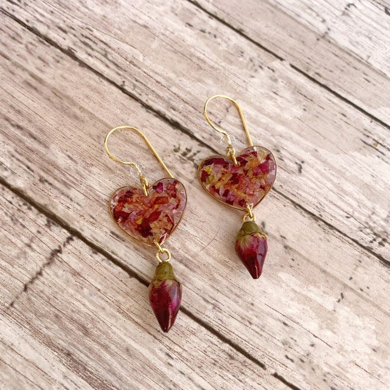 Puede incluir: Un par de pendientes de coraz&oacute;n de oro con un colgante de capullo de rosa rojo. Los corazones est&aacute;n llenos de p&eacute;talos de rosa prensados.