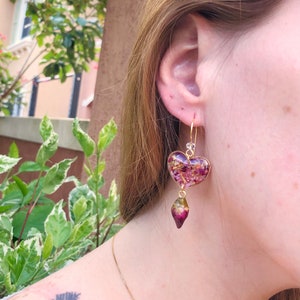 Puede incluir: Pendientes de coraz&oacute;n de oro con resina morada y rosa llena de flores secas y un colgante de l&aacute;grima morado y verde.
