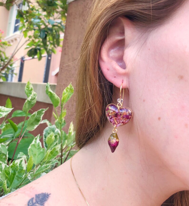 Puede incluir: Pendientes de coraz&oacute;n de oro con resina morada y rosa llena de flores secas y un colgante de l&aacute;grima morado y verde.