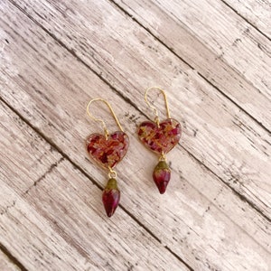 Puede incluir: Un par de pendientes de coraz&oacute;n dorados con p&eacute;talos de rosa secos encapsulados en resina transparente. Cada pendiente tiene un encanto de capullo de rosa que cuelga.