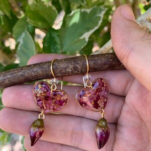 Puede incluir: Un par de pendientes de coraz&oacute;n dorados con p&eacute;talos de flores secas encapsulados en resina. Cada pendiente tiene un colgante en forma de l&aacute;grima con un dise&ntilde;o similar.