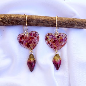 Puede incluir: Un par de pendientes de coraz&oacute;n de oro con un colgante de capullo de rosa. Los corazones est&aacute;n llenos de p&eacute;talos de flores rosas y blancas prensados.