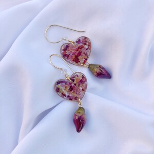 Puede incluir: Un par de pendientes de coraz&oacute;n de oro con un dise&ntilde;o floral rosa y blanco. Cada pendiente tiene una rosa rosa colgante.
