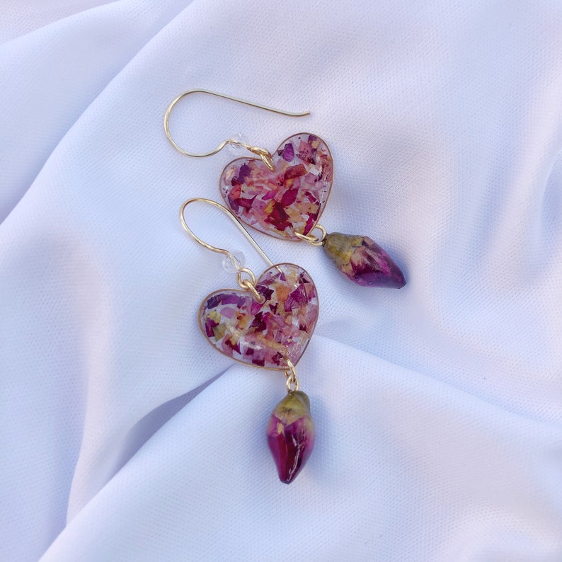 Puede incluir: Un par de pendientes de coraz&oacute;n de oro con un dise&ntilde;o floral rosa y blanco. Cada pendiente tiene una rosa rosa colgante.