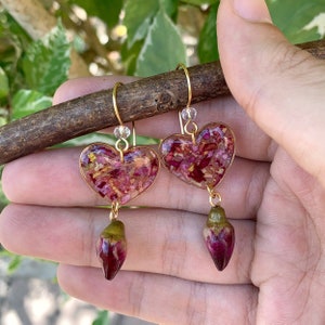Puede incluir: Un par de pendientes de coraz&oacute;n dorados con p&eacute;talos de flores secas rosas y rojos encapsulados en resina transparente. Cada pendiente tiene una yema de flor rosa y verde colgante.