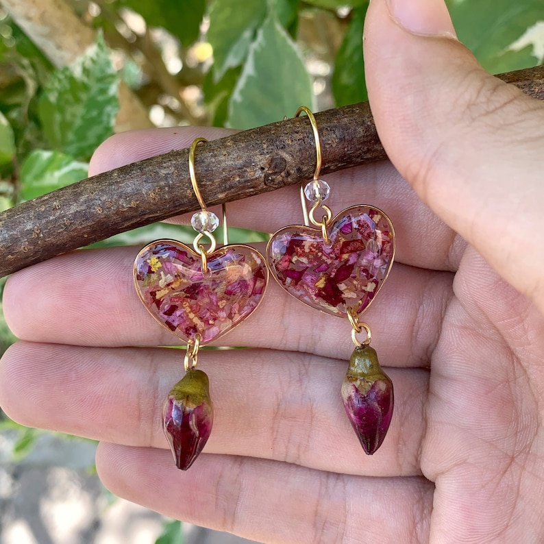 Puede incluir: Un par de pendientes de coraz&oacute;n dorados con p&eacute;talos de flores secas rosas y rojos encapsulados en resina transparente. Cada pendiente tiene una yema de flor rosa y verde colgante.