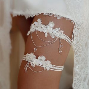 May include: White bridal garter with delicate lace flowers and crystal embellishments. The garter has a satin ribbon and a silver chain with crystal drops.