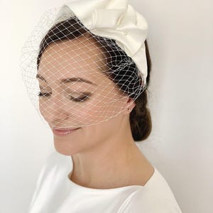 Pode incluir: Uma tiara de cetim branco com um laço grande e um véu de rede fina. O laço é adornado com uma pequena pérola. O véu é uma rede delicada, e a tiara é usada na cabeça. Ideal para casamentos.