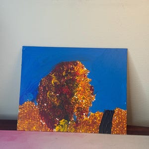 May include: An abstract painting of a tree with a blue sky background. The tree is painted with shades of orange, yellow, and red, with a dark brown trunk. The painting is on a rectangular canvas.