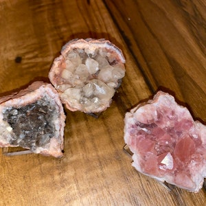 Könnte beinhalten: Drei Geoden-Exemplare mit unterschiedlichen Kristallformationen. Die erste Geode hat klare Quarzkristalle, die zweite hat dunkelgraue Kristalle und die dritte hat rosa Amethystkristalle.