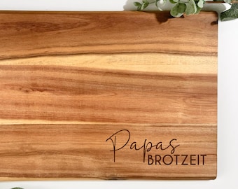 Pode incluir: Tábua de corte retangular de madeira castanha clara com o texto "Papas Brotzeit" gravado. A madeira apresenta veios visíveis. Folhas verdes dispostas ao redor da tábua.