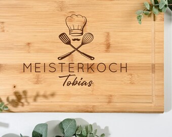 Pode incluir: Tábua de corte retangular de bambu com ranhura para suco. A tábua apresenta um chapéu de chef, utensílios cruzados e as palavras "MEISTERKOCH Tobias" gravadas. A vegetação envolve a tábua.