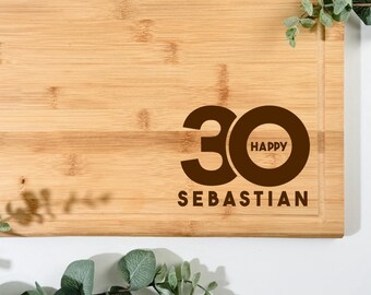 Pode incluir: Uma tábua de corte retangular de bambu com ranhura para sumo. A tábua apresenta o texto "30 HAPPY SEBASTIAN" em castanho. O texto "GRAFIK 1" está no canto superior esquerdo. A vegetação envolve a tábua.