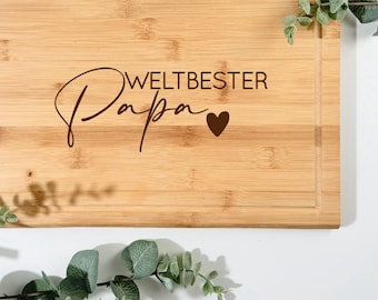 Pode incluir: Tábua de corte retangular de bambu com ranhura para sumo. As palavras "WELTBESTER Papa" e um coração estão gravadas na madeira. A vegetação é disposta ao redor da tábua sobre um fundo branco.