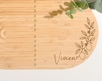 Pode incluir: Uma tábua de servir oval de bambu com um padrão de arco esculpido no lado esquerdo e uma ilustração botânica gravada a laser de limões com folhas à direita. O nome Vivien está gravado em letra cursiva abaixo do desenho do ramo.