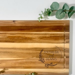 May include: A rectangular wooden cutting board with a juice groove. The board has a light brown color with visible wood grain. The text "MATHILDA und DENNIS" is engraved within a floral wreath. The text "GRAFIK I" is in the upper left corner.