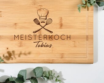 Pode incluir: Uma tábua de corte retangular de bambu com um chapéu de chef, utensílios cruzados e o texto gravado "MEISTERKOCH Tobias". A tábua tem uma borda rebaixada e é rodeada por vegetação.