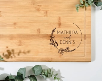 Pode incluir: Tábua de corte retangular de bambu com borda entalhada. A tábua apresenta o texto "MATHILDA und DENNIS" dentro de um desenho de coroa floral. O texto "GRAFIK 1" está no canto superior esquerdo.