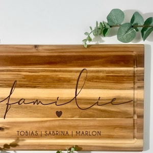 Könnte beinhalten: Rechteckiges Holzschneidebrett mit Saftrille. Das Wort "familie" ist in der Mitte geschrieben, darunter ein Herzsymbol. Die Namen "TOBIAS | SABRINA | MARLON" stehen unter dem Herzen. Der Text "GRAFIK 1" befindet sich oben links.