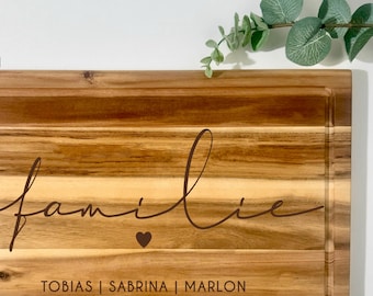 Pode incluir: Tábua de corte retangular de madeira com ranhura para suco. A palavra "familie" está escrita no centro, com um símbolo de coração abaixo. Os nomes "TOBIAS | SABRINA | MARLON" estão abaixo do coração. O texto "GRAFIK 1" está no canto superior esquerdo.