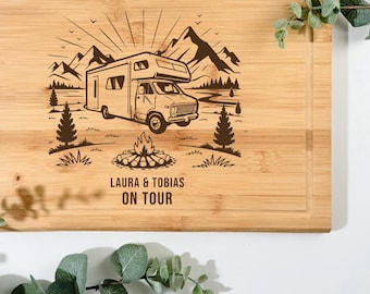 Pode incluir: Tábua de corte retangular de bambu com um design entalhado. O design apresenta uma autocaravana, montanhas, árvores e uma fogueira. O texto "LAURA & TOBIAS ON TOUR" está abaixo. A tábua tem um sulco para suco na borda.