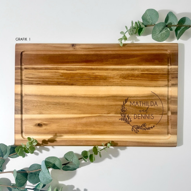 May include: A rectangular wooden cutting board with a juice groove. The board has a light brown color with visible wood grain. The text "MATHILDA und DENNIS" is engraved within a floral wreath. The text "GRAFIK I" is in the upper left corner.