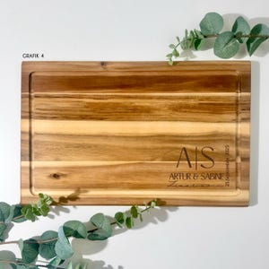 May include: A rectangular wooden cutting board with a juice groove. The board is engraved with the initials "AIS", the names "ARTUR & SABINE", and the date "23. September 2025". The text "GRAFIK 4" is in the upper left corner.