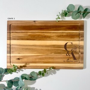 May include: A rectangular wooden cutting board with a juice groove. The board features the initials "C & A" with floral accents and the date "25. Oktober 2025" engraved. The text "GRAFIK 3" is in the upper left corner.