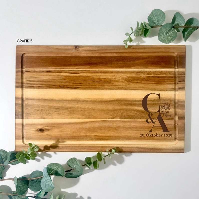 May include: A rectangular wooden cutting board with a juice groove. The board features the initials "C & A" with floral accents and the date "25. Oktober 2025" engraved. The text "GRAFIK 3" is in the upper left corner.