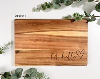 Pode incluir: Uma tábua de corte retangular de madeira com um acabamento de grão natural. O nome "Michelle" está gravado em itálico, acompanhado por um símbolo de coração e a frase "SCHÖN, DASS ES DICH GIBT".