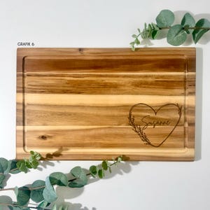 May include: A rectangular wooden cutting board with a juice groove. The board features a heart-shaped design with the name "Susanne" engraved inside. The text "GRAFIK 6" is in the upper left corner. Greenery is placed around the board.
