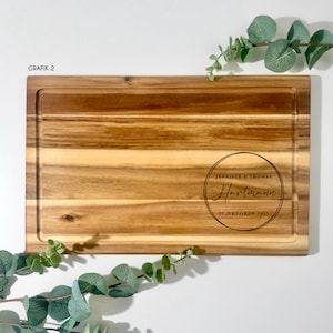 May include: A rectangular wooden cutting board with a juice groove. The board features engraved text within a circle: "JENNIFER & THOMAS Hartmann 21.OKTOBER 2023". The board is surrounded by green foliage. The text "GRAFIK 2" is in the upper left corner.
