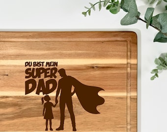 Pode incluir: Tábua de corte de madeira com um design entalhado. O design apresenta as palavras "DU BIST MEIN SUPER DAD DEINE SINA" e uma silhueta de um pai e uma criança. A tábua é retangular com uma ranhura para sumo.