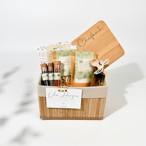 Könnte beinhalten: Ein Bambus-Geschenkkorb gefüllt mit Gourmet-Lebensmitteln, darunter Gewürze, Salz, Grissini und Olivenöl. Der Korb ist mit einem Holzbrett, das mit dem Wort "Chefkoch" graviert ist, und einer Karte mit der Aufschrift "Von Herzen" verziert.