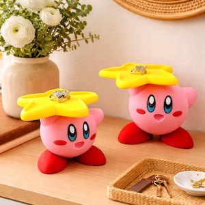 Puede incluir: Dos sujetadores de anillos rosas con forma de Kirby con tapas amarillas en forma de estrella y pies rojos. Cada soporte tiene un anillo en la parte superior. El fondo incluye un jarrón de flores blancas, un espejo y una pequeña bandeja con llaves.