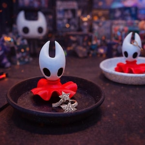 Puede incluir: Una figura blanca y roja de un personaje del videojuego Hollow Knight, colocada en un plato negro con un anillo en forma de estrella. Otra figura está en un plato blanco. Otras figuras están en el fondo.