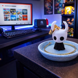 Puede incluir: Una figura blanca y negra con cuernos curvos, sosteniendo un anillo dorado, se encuentra en un plato moteado azul claro. Una pulsera dorada rodea el plato. Una configuración de juego con un monitor y un teclado está en el fondo.