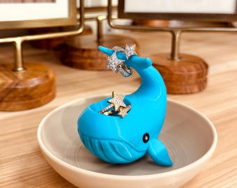 May include: A turquoise whale-shaped ring holder with a small dish base. The whale holds several rings with star and diamond designs. The holder sits on a light wood surface, with framed pictures in the background.