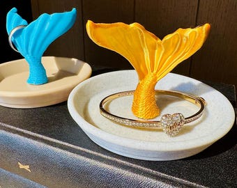 May include: Two mermaid tail ring holders, one blue and one yellow, each atop a small dish. A gold bracelet with a heart-shaped charm rests in the yellow holder. The holders sit on a stack of dark books.