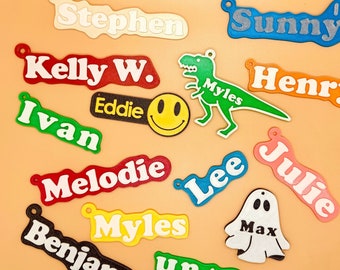 Op de afbeelding: Een verzameling kleurrijke, gepersonaliseerde naamstickers met verschillende ontwerpen, waaronder een smiley, een dinosaurus en een spook. De namen zijn Stephen, Sunny, Kelly W., Eddie, Ivan, Melodie, Lee, Julie, Myles, Benjamin en Henry.
